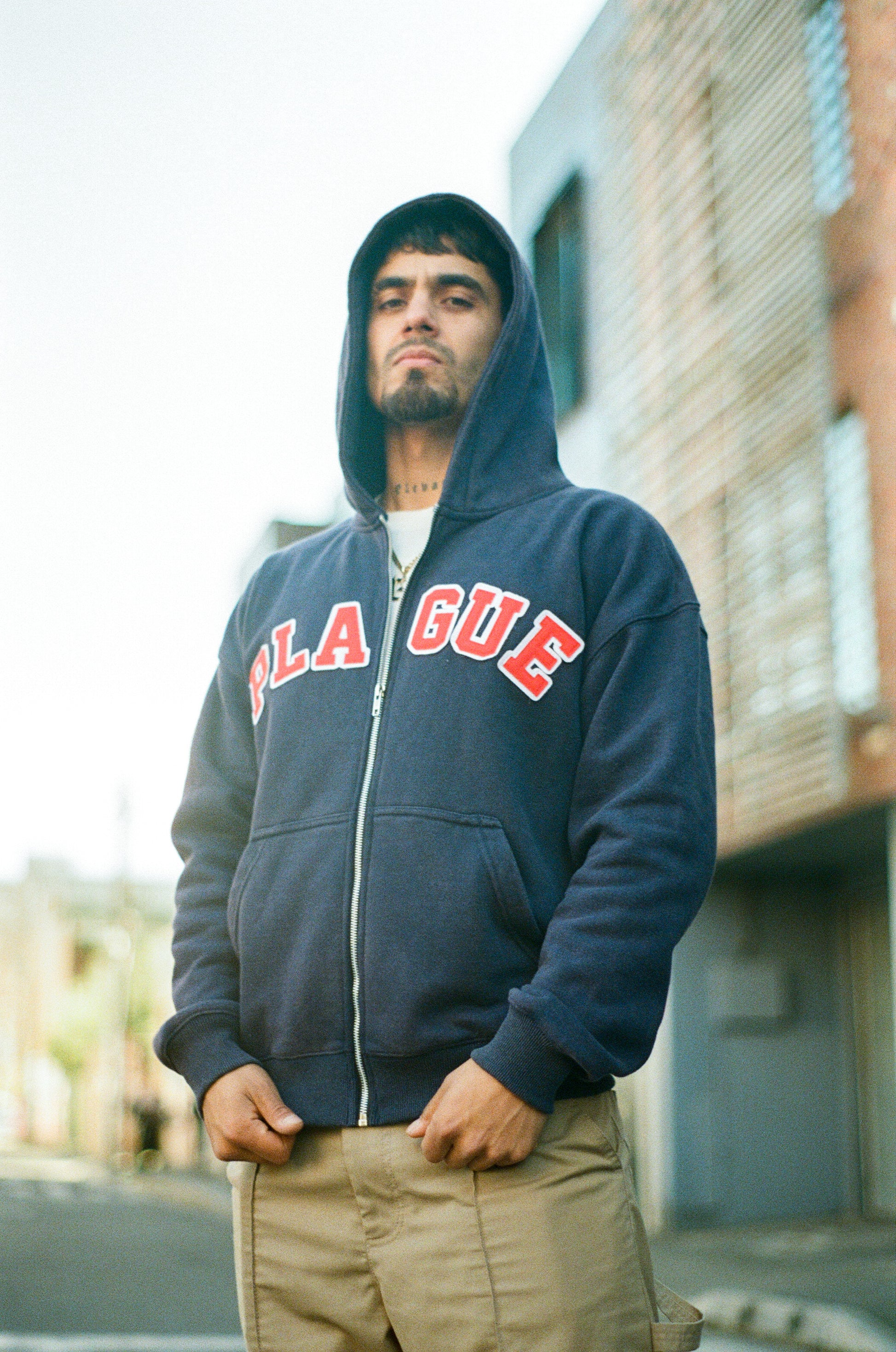 Man wearing a navy blue hoodie with 'PLAGUE' text, standing outdoors.