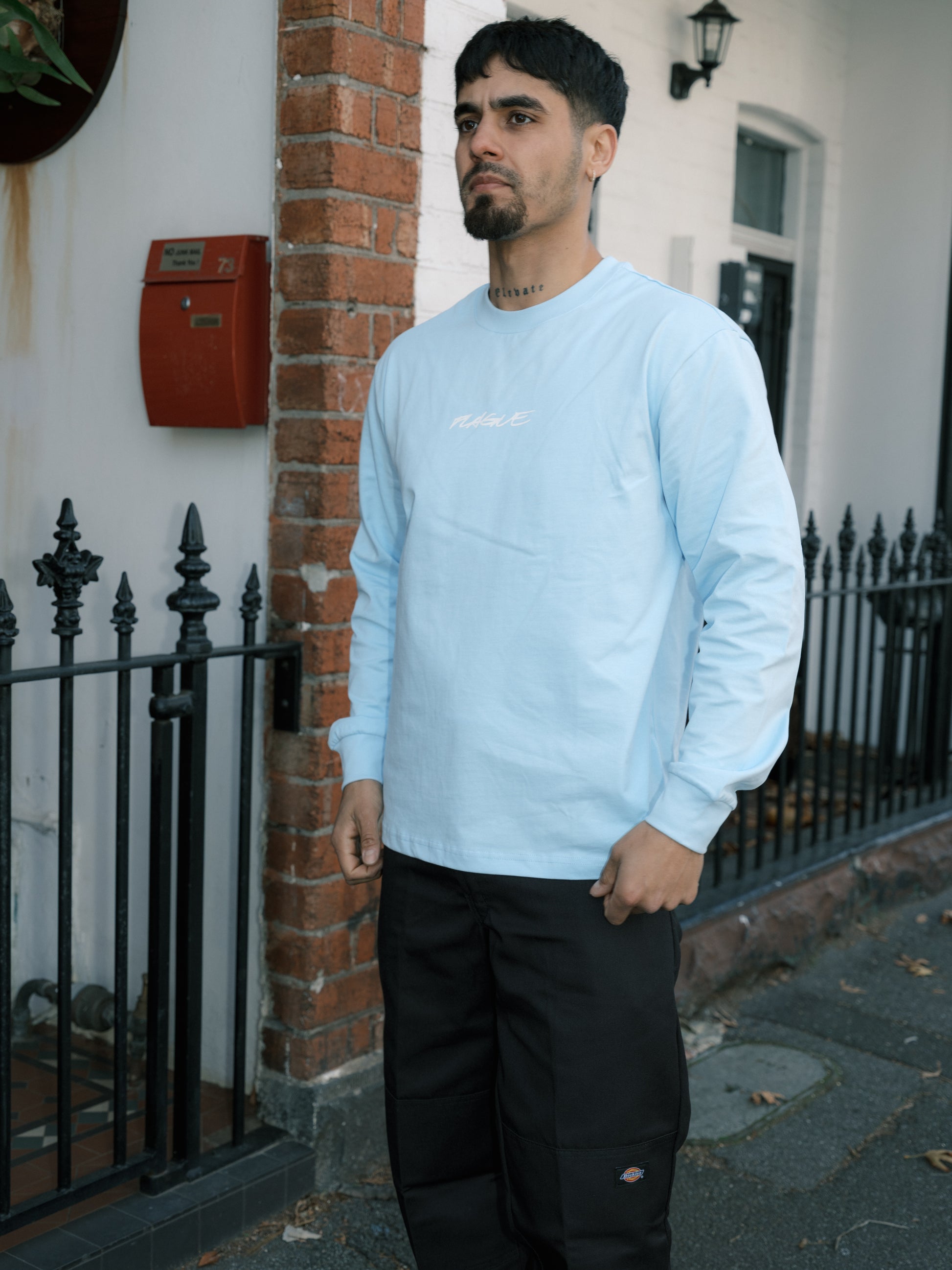 Man wearing a light blue long sleeve tee standing outside a building with a brick wall and black metal gate.