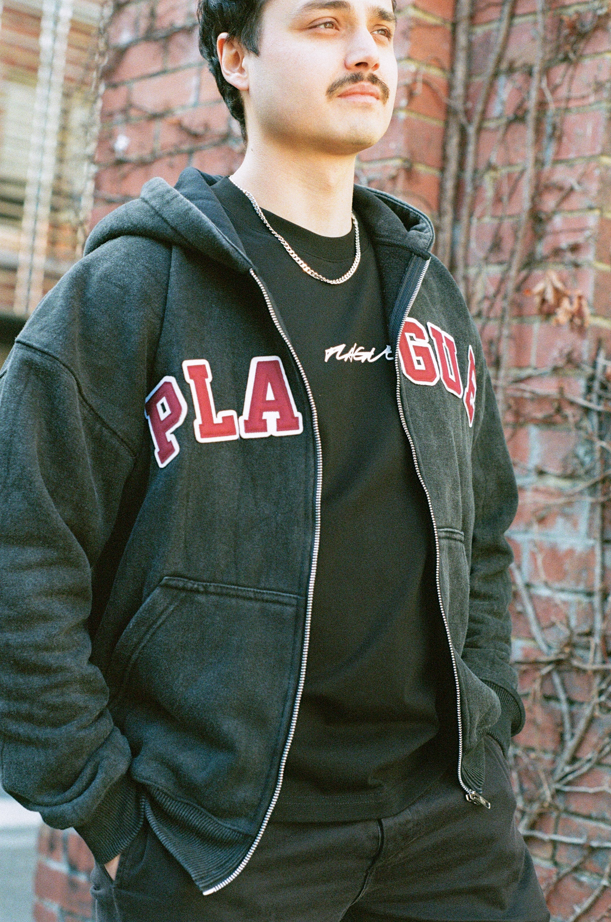 Person wearing a dark hoodie with 'PLAGUE' text, standing against a brick wall.